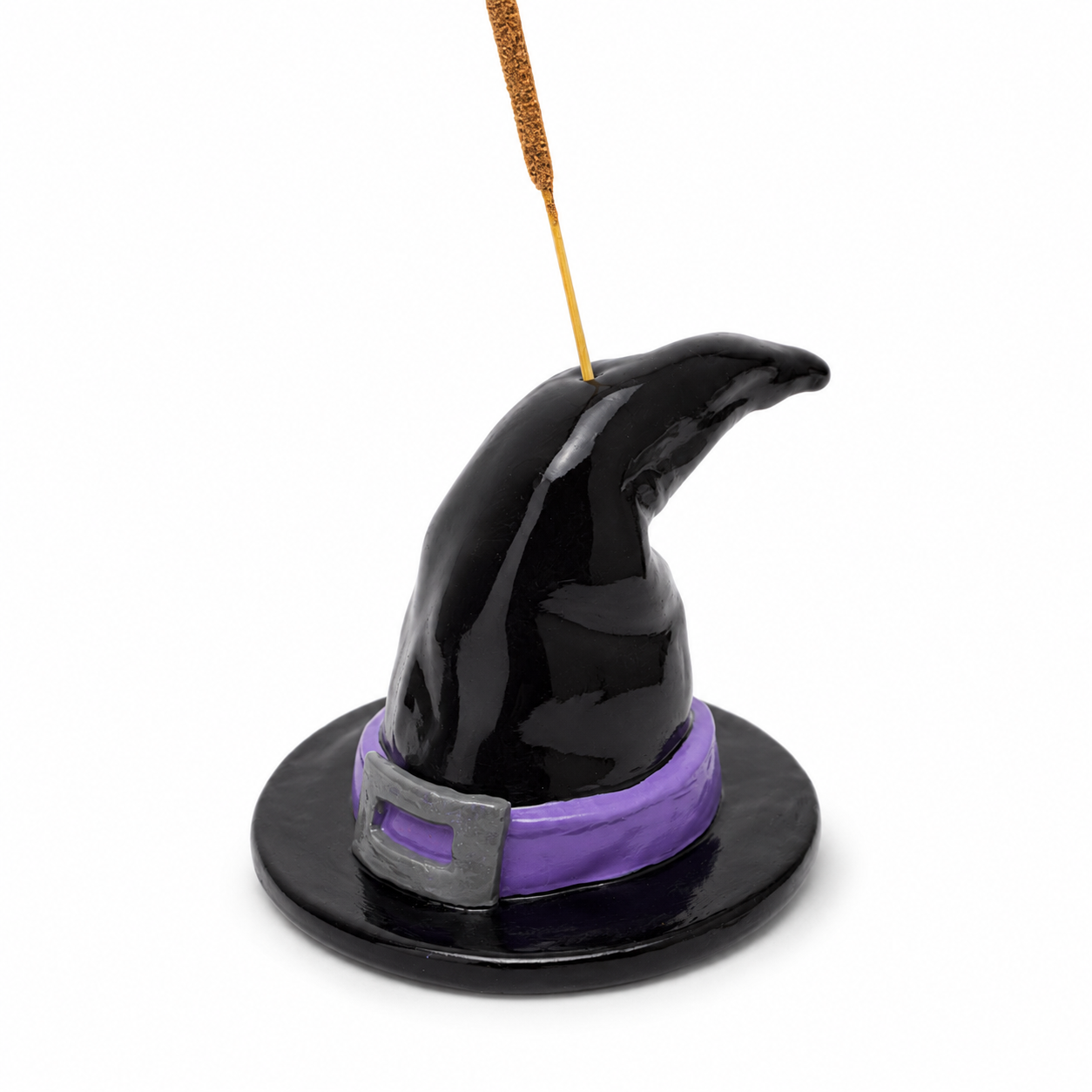 Incensário em formato de chapéu de bruxa preto, com faixa roxa, fivela prateada e incenso inserido na parte superior.